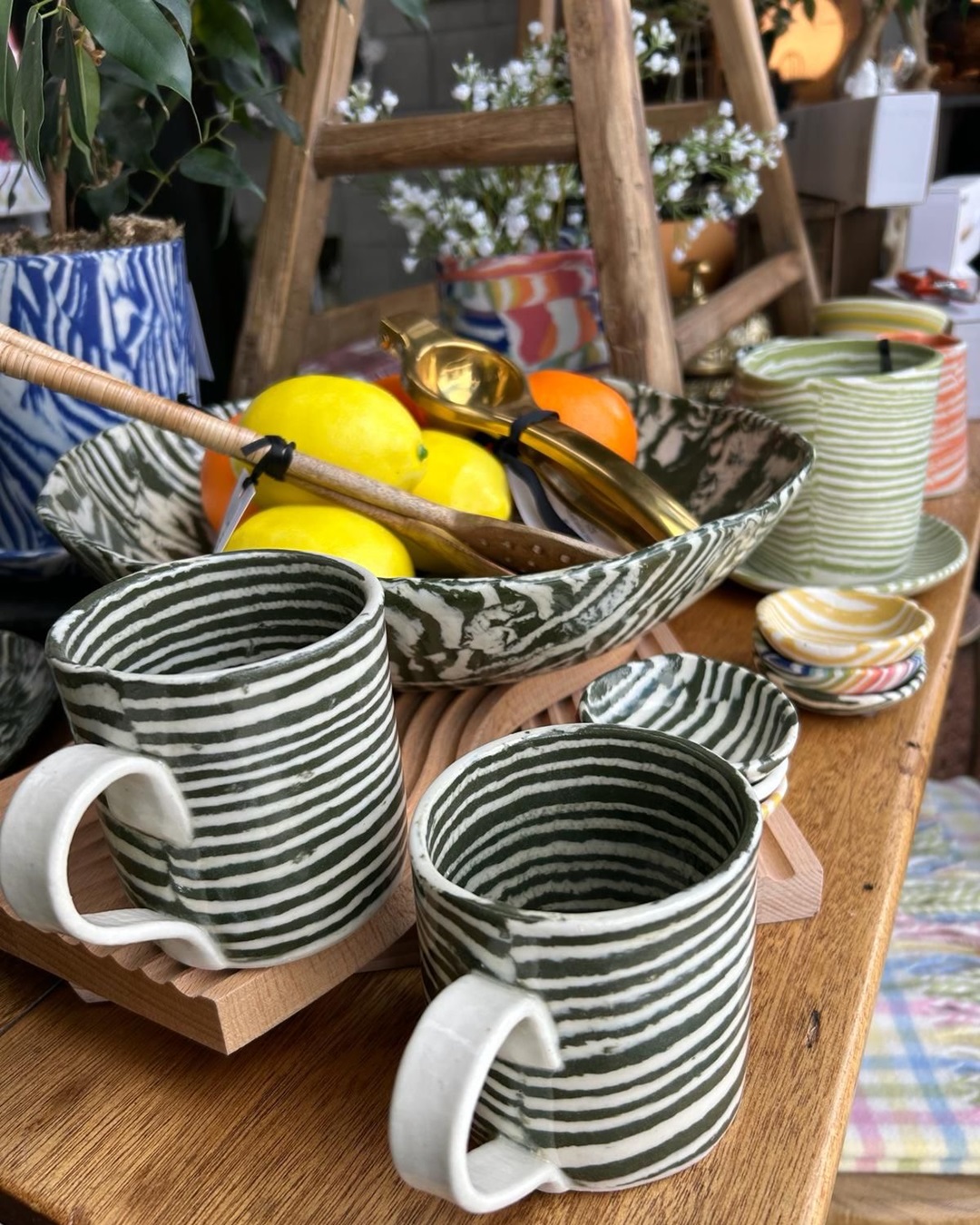 Green striped serving bowl with serving spoons and fruit surrounded by cups, planters and bowls