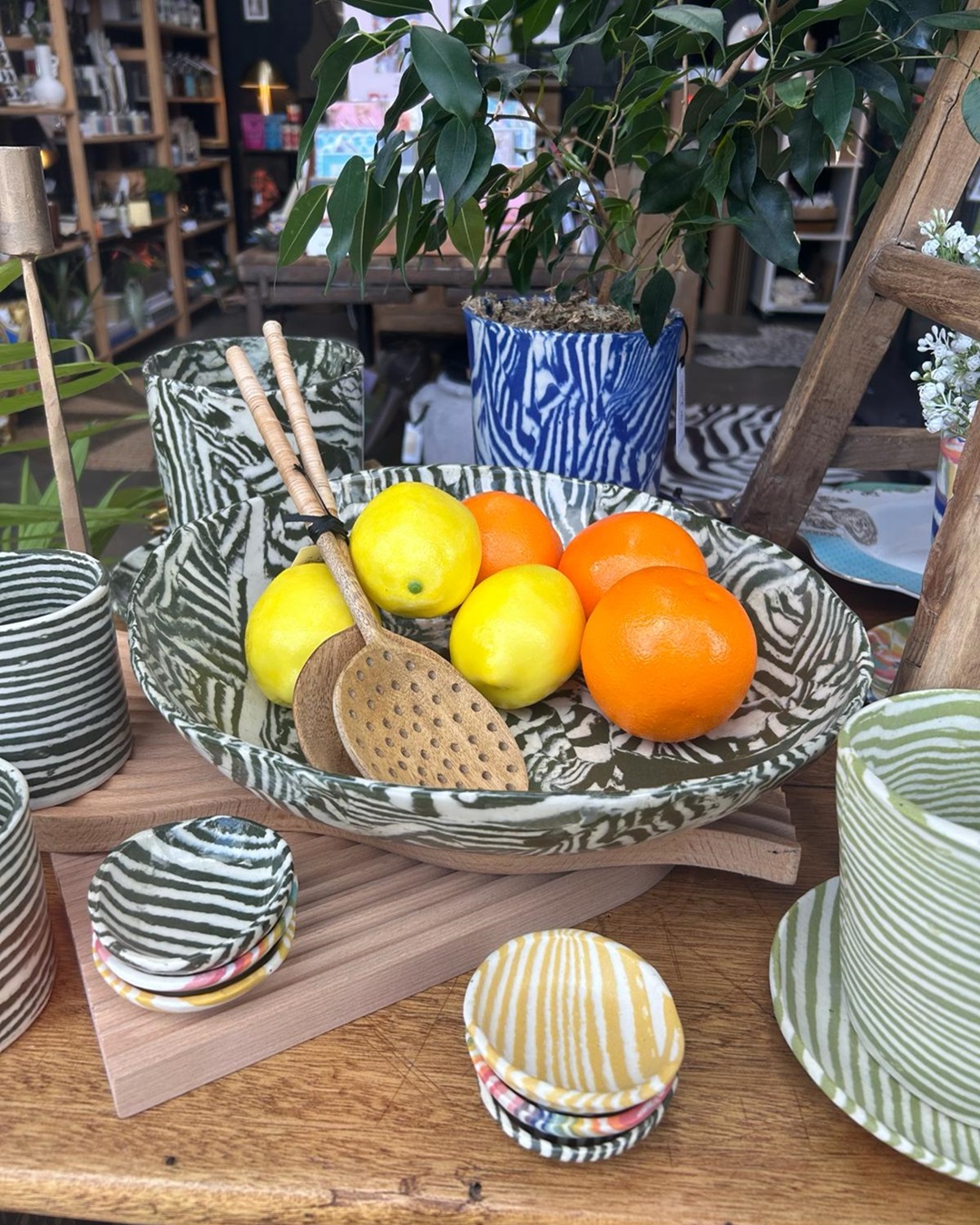 Green striped serving bowl with serving spoons and fruit surrounded by cups, planters and bowls